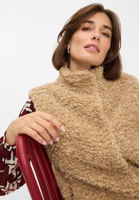 Beige vest met krullend textuur en hoge kraag. Model steunt hand op een rode stoel. Patroon blouse is zichtbaar onder het vest. Neutrale achtergrond.
