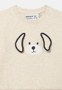 Ανοικτό μπεζ φούτερ με γραφικό πρόσωπο σκύλου. Διαθέτει ναυτικό περίγραμμα για τα αυτιά, μαύρα μάτια και μύτη. Απαλό ύφασμα με ριμπώ γιακά.