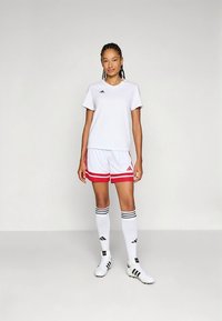 Fehér adidas sportpóló rövid ujjú, fehér rövidnadrággal, amely piros csíkokkal, fekete betétekkel és térdig érő csíkos zoknival van párosítva.