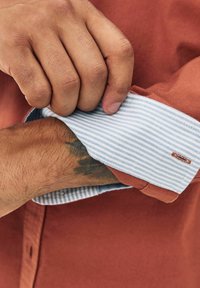 Rust-colored shirt with a blue and white striped cuff. The cuff features a small logo accent near the edge. Cotton fabric with a smooth texture.