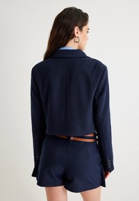 Granatowa krótka marynarka z teksturowanej tkaniny, z kołnierzem w kształcie lapelu i brązowym paskiem w talii, stylizowana na szorty.