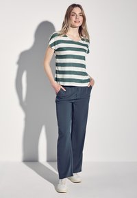 Maglietta a maniche corte a righe verde e bianca, abbinata a pantaloni larghi grigio scuro e sneakers bianche, con le mani nelle tasche.