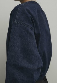 Tummansininen farkkukangascollege, jossa rennosti laskeutuva istuvuus, pyöreä pääntie ja lasketut olkapäät, korostaen tekstuurillista kangasta ja näkyviä saumoja.