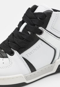 Hvite og svarte høye sneakers med skinnoverdel, teksturerte svarte lisser, preget logo og gummisåle med grå aksent.