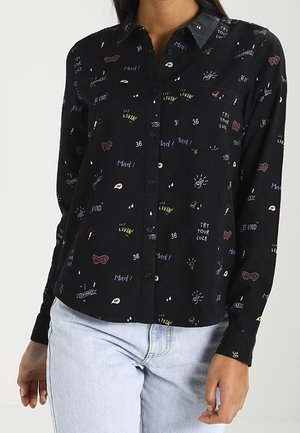 Schwarzes Hemd mit Knopfleiste, bunten kleinen grafischen Drucken und Text, kombiniert mit hellblauen Jeans.