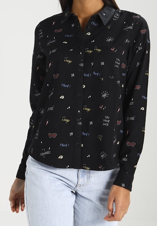 Chemise noire boutonnée avec de petits imprimés graphiques colorés et du texte, portée avec un jean bleu clair.