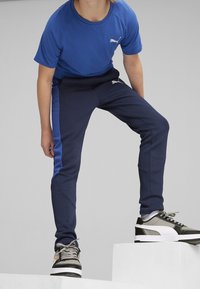 Blaues kurzärmliges Hemd und marineblaue Jogginghosen mit blauen Seitenstreifen. Auffälliges PUMA-Logo auf dem Hemd und den Hosen. Graue Sneakers mit schwarzen Akzenten.