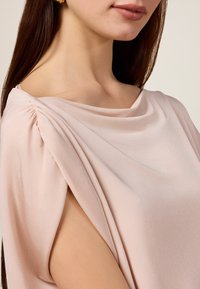 Blusa drappeggiata di un delicato rosa con dettagli arricciati alla spalla, che mette in mostra una texture morbida e una vestibilità rilassata. Design con maniche aperte che rivela la pelle.