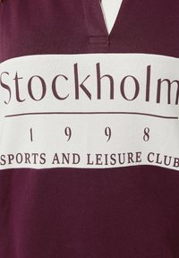 Marinröd och vit pikétröja i bomull. Har en v-ringad hals och med "Stockholm 1998 Sports and Leisure Club" tryckt i fet stil.