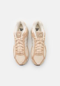 Baskets avec une tige en maille beige et des accents crème, trois rayures blanches, et une semelle en caoutchouc texturé. Logo affiché sur la languette.