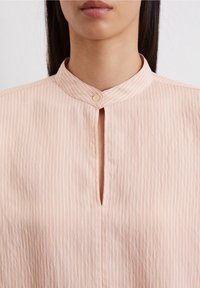 Blouse rose clair avec des rayures verticales, col montant et une patte de boutonnage à l'avant avec un seul bouton. Texture lisse et minimalisme dans les détails.