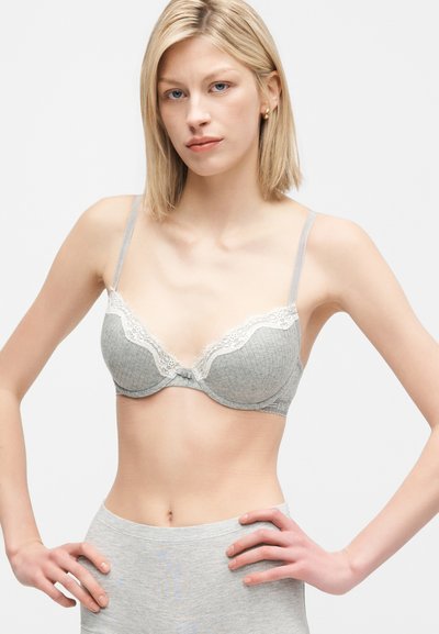 Lyshåret kvinde med blå øjne iført en grå blondekantet bh og matchende shorts med taljebånd, stående med hænder på hofterne foran en ensfarvet baggrund.