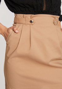Beige přizpůsobená sukně s sklady, vypasovaným pasem a páskem se zlatým kováním. Hladká textura a boční kapsy jsou viditelné.