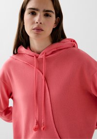 Roze hoodie van zacht materiaal, met een losse fit, zijnaad en contrasterende trekkerkoordjes met textuur aan de uiteinden.