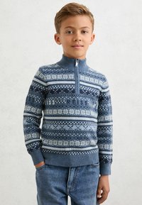 Plavi pleteni pulover s visokim ovratnikom i zatvaračem. Ima razne plave i bijele uzorke, uključujući pahuljice i pruge. Uparen s plavim trapericama.