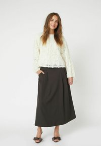 Weiße strukturierte Bluse mit langen Ärmeln, kombiniert mit einem dunkelgrauen, knielangen Rock mit Falten und seitlichen Taschen. Sandalen mit schwarzen Schleifenverzierungen.