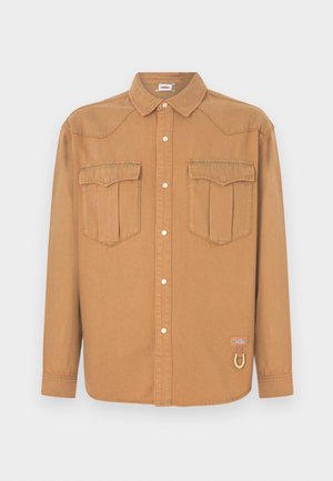 Hnedá denimová košeľa s dvoma náprsnými vreckami, špicatým golierom, zapínaním na gombíky vpredu a kontrastným prešitím. Má logo nášivku a detail s pútkom.