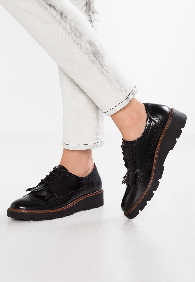 Chaussures brogue en cuir noir avec détail à franges, portées avec un pantalon blanc, montrées avec les jambes croisées sur un fond uni.