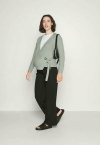 Cardigã tricotado verde-claro com um design de envoltório e laço na cintura, combinado com calças largas pretas e sandálias pretas.