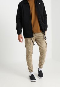 Czarna kurtka z miękkim kołnierzem, brązowa koszula pod spodem, spodenki cargo w kamuflażu i czarne buty na slip-on z białymi podeszwami oraz skarpetki za kostkę.