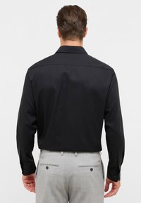 Camicia nera a maniche lunghe con colletto classico e polsini con bottoni, abbinata a pantaloni grigi chiari con tasche posteriori e dettagli con bottoni.