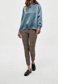 Satinblå långärmad topp med hög krage, tillsammans med bruna byxor i kockrutor i slim fit och svarta loafers.