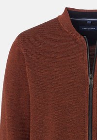 Zippertröja i strukturerat rostfärgat stickat material. Har en ståkrage och svart dragkedja. Märkesetikett synlig vid halslinningen.