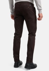 Pantalones ajustados de color marrón oscuro, hechos de una tela suave, con bolsillos traseros con botones y llevados con zapatos negros.
