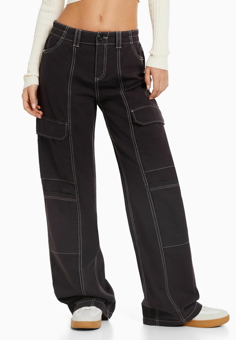Femme portant un pantalon cargo noir à jambes larges avec des surpiqûres blanches contrastantes, associé à des baskets blanches et un pull court côtelé crème.
