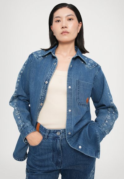Cămașă din denim cu un finisaj albastru estompat, nasturi cu prindere tip snaps, guler ascuțit și buzunar pe piept cu un accent din piele maro pe lateral.