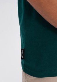 Tmavě zelené bavlněné tričko s božním štítkem s nápisem "LEVEL UP". Krátké rukávy, kulatý výstřih a jednoduchý, texturovaný povrch.