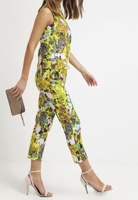Completo floreale a due pezzi con top senza maniche e pantaloni cropped in giallo, verde e grigio con una finitura lucida. Accompagnato da tacchi nudi.
