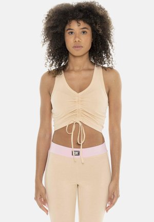 Femme aux cheveux bouclés portant un crop top beige froncé et des leggings assortis avec une ceinture rose, debout sur fond blanc.