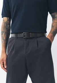 Zwarte leren riem met een rechthoekige metalen gesp, gedragen over donkergrijze broek. De outfit bestaat uit een donkerblauw polohemd.