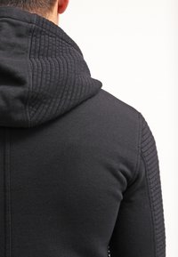 Černá mikina s huňatou texturou na ramenou a kapuci, vyrobená z hladkého materiálu s vysokým výstřihem.