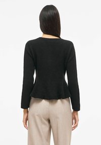 Melnas pepluma džemperis ar garām piedurknēm, teksturētu virsmu, apaļu kakla izgriezumu un mīkstu audumu, valkāts kopā ar bēšām biksēm. Vienkāršs fons.