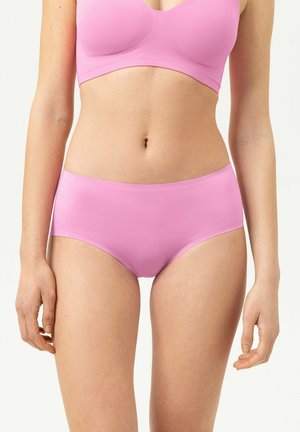Femme portant un soutien-gorge rose clair assorti à une culotte taille haute, visible de la poitrine jusqu'aux cuisses sur un fond uni.
