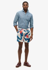 Camisa de mezclilla abotonada, pantalones cortos de baño con estampado de hojas tropicales y zapatos de barco de cuero burdeos con cordones. Atuendo casual de verano.