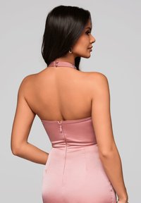 Een vrouw staat met haar rug naar ons toe, gekleed in een roze halterjurk met een ritsdetail, waardoor haar lange donkere haar en profiel zichtbaar zijn.