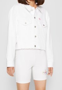 Valkoinen denimpitkä takki, jossa on painonapit ja rintataskut, sekä kevyttä kulumista helmassa. Valkoiset shortsit, joissa on värikkäitä graafisia yksityiskohtia.