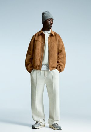 Brun ruskinds bomberjakke, hvid hættetrøje, lysegrå sweatpants og grå sneakers. Modellen bærer en grå strikbeanie. Minimalistisk baggrund.