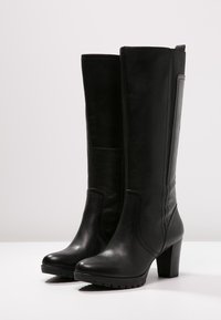 Botas de cuero negras hasta la rodilla con tacones cuadrados y puntas redondeadas, que cuentan con paneles laterales elásticos y detalles de costura, apoyadas sobre una superficie blanca.