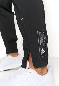 Czarne spodnie sportowe z zamkiem na kostce, pomarańczowymi przeszyciami i białą metką z logo Adidas. W zestawie z białymi butami.