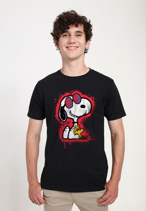 Ung mand smilende iført sort t-shirt med Snoopy og Woodstock grafik og rød malingstænk baggrund foran.