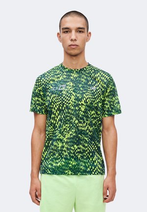 Homme portant un t-shirt de sport à manches courtes vert et noir à motif géométrique et un short vert clair, debout devant un fond uni.