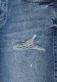 Tissu en denim bleu avec une déchirure effilochée horizontale exposant des fils blancs, près d'une poche avec des coutures visibles et une rivure.