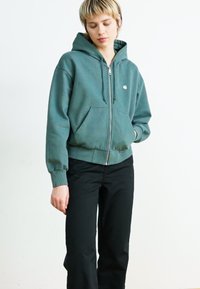 Grønn zip-up hettegenser i mykt materiale med frontlommer, snøring i hetten og ribbede mansjetter, parret med svarte bukser. Enkelt design og passform.
