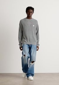 Mann, der einen grauen Pullover, zerrissene blaue Jeans und weiße Sneaker trägt, vor einem schlichten weißen Hintergrund geht.
