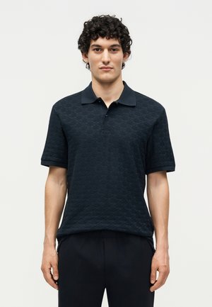 Νέος άνδρας με κατσαρά μαλλιά, που φοράει μαύρο πικέ πόλο με κοντά μανίκια και μαύρο παντελόνι, στέκεται μπροστά σε ένα απλό φωτεινό φόντο.