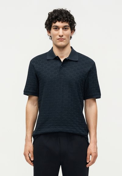 Giovane uomo con capelli ricci che indossa una polo nera texturizzata a maniche corte e pantaloni neri, in piedi davanti a uno sfondo chiaro e semplice.